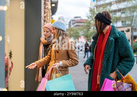 Three diverse young women enthusiastically window shopping in winter city. Friends of different ethnicities carrying colorful bags, looking at festive Stock Photo