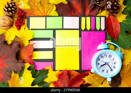 Bright sticky notes and an alarm clock are arranged among colorful autumn leaves, creating a lively fall display. Stock Photo