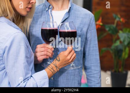 An unrecognizable young couple makes a toast while drinking red wine with a glass in hand against the backdrop of an outdoor wooden house Stock Photo