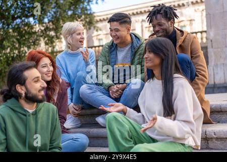 Diverse group of young students relaxing and socializing on campus steps. Multicultural friends using smartphones, chatting, and enjoying time togethe Stock Photo