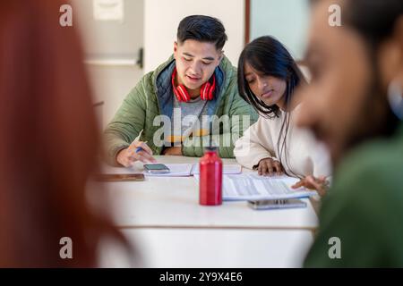 Asian college students working on academic project in university library. Diverse friends using smartphone and notes for collaborative study. Concept Stock Photo