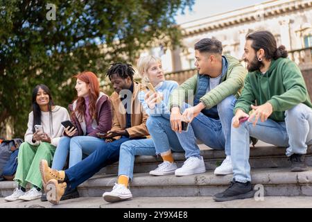 Diverse group of young students relaxing and socializing on campus steps. Multicultural friends using smartphones, chatting, and enjoying time togethe Stock Photo