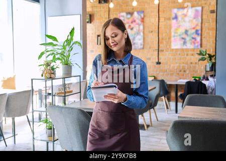 Young woman worker, owner in apron holding notepad pen in restaurant, coffee shop Stock Photo
