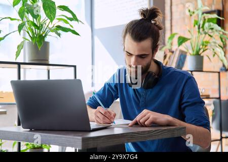 Young male university student studying using laptop, writing in notebook Stock Photo