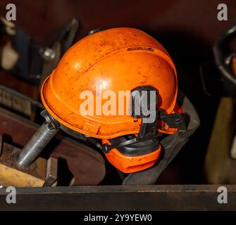 Photograph of an old and dirty orange safety hard hat with hearing protection laying on a metal table in a workshop. Stock Photo