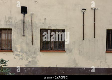 Old tools hanging on a weathered building wall with barred windows. Concept of rural lifestyle, gardening equipment and vintage architecture Stock Photo