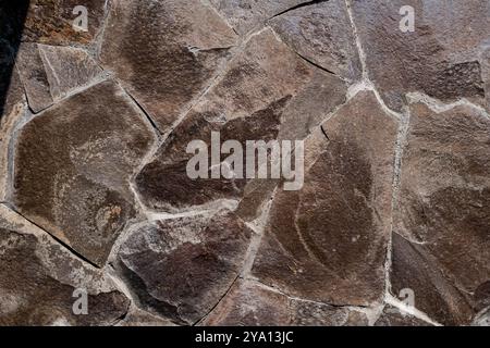 A close-up view of a textured stone wall featuring irregularly shaped brown stones with visible cracks and variations in color. The surface appears na Stock Photo