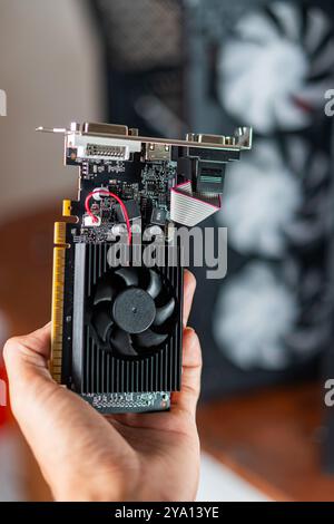 A person holding a graphics card with a black cooling fan and various connectors. The background shows a computer case with visible fans, indicating a Stock Photo