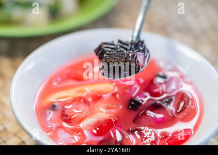 A close-up of a bowl filled with colorful dessert, featuring jelly cubes and various fruits in a sweet syrup. A spoon is lifting a piece of black jell Stock Photo