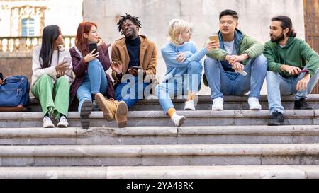 Group of diverse young students sitting on university steps, using smartphones and socializing. Multicultural friends enjoying time together on campus Stock Photo