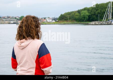 A young brunette woman stands with her back to the camera, gazing at a serene seascape with a peaceful island with lush green vegetation. Nature conne Stock Photo