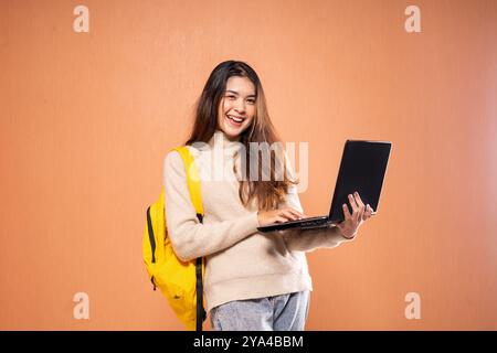 Cheerful Student Using Laptop with Yellow Backpack Against Orange Wall Stock Photo