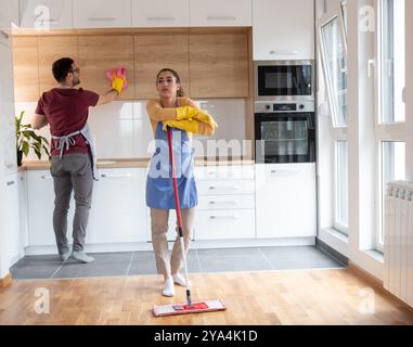 Young couple cleaning their modern kitchen together while wearing aprons on sunny day Stock Photo
