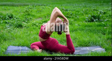 Young beautiful brunette haired woman performs the complicated yoga posture of the dove or Eka pada rajakapotasana on her mat in the middle of nature Stock Photo