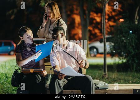 Group of professionals collaborating on project outdoors during evening, discussing documents and using digital technology on a park bench. Stock Photo
