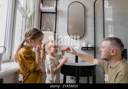 A father teaches his two young daughters how to brush their teeth in the bathroom. Stock Photo