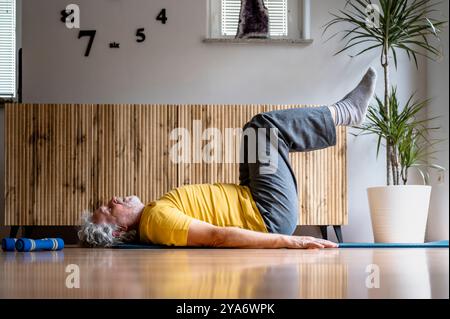 Profile view of a senior man in bright yellow tshirt lying on his back with his legs lifted up. Exercising pilates for strong core muscles for a healt Stock Photo