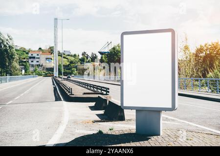 A blank advertising billboard mockup stands prominently on a roadside near a bridge. The road is empty, bordered by safety rails, trees, and distant h Stock Photo