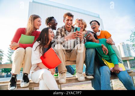 Group of diverse students sitting outdoors, laughing and using a cellphone together, enjoying social media, connecting through technology while holding notebooks and sharing moments on campus. High quality photo Stock Photo