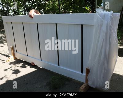unpacking a light polyethylene door leaf using stands in the courtyard of a private house, a man removes plastic packaging material from a white door Stock Photo
