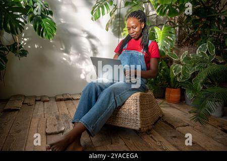 Smiling African American woman using laptop surrounded by plants and flowers. Flower shop owner Stock Photo