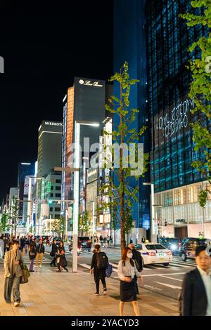 View along the brightly lit Ginza shopping street at night with the Chanel building and various others on the street. Pavement busy, people walking. Stock Photo