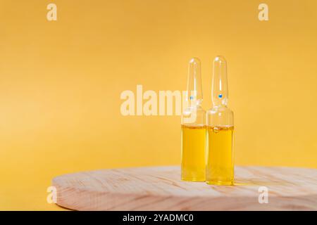 Two glass ampoules containing yellow liquid, placed on a wooden surface with a soft yellow background. The ampoules have a blue dot on the top, sugges Stock Photo