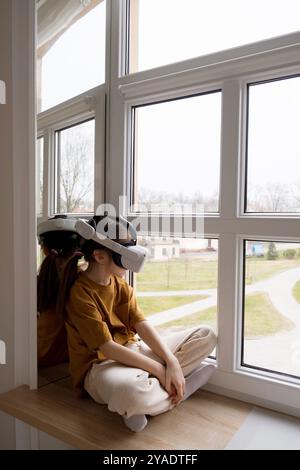 A young girl, wearing a VR headset, sits by a window, gazing out at the real world. Stock Photo