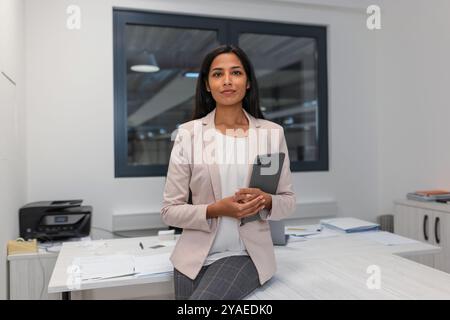 Smiling Indian businesswoman posing in a company office, holding a tablet and looking at the camera. Business and managers concepts. Stock Photo