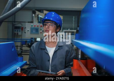 Indian woman engineer with a blue helmet holding a tablet while checking and controlling working machine parts and tools. Stock Photo