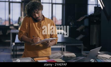 Arabian Indian male business man working busy interior designer talking with client online discuss on mobile phone fabric tissue samples write in Stock Photo