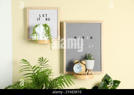 Letter boards with phrases Let It Be, Never Give Up, alarm clock and plants on beige wall Stock Photo
