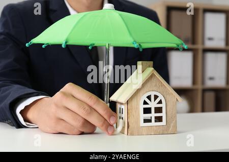 Real estate insurance. Man holding small umbrella above wooden house figure at light table, closeup Stock Photo