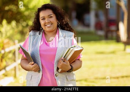 Happy student in a pink shirt holding books and a tablet outdoors Stock Photo