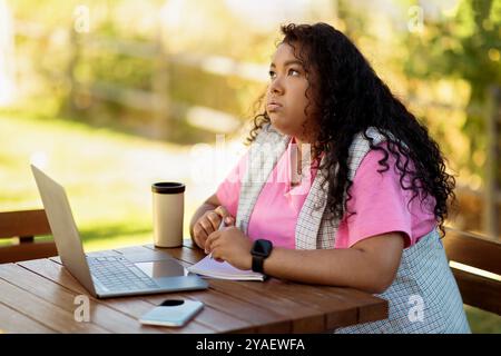 Young woman pondering while working on laptop at outdoor cafe in daylight Stock Photo