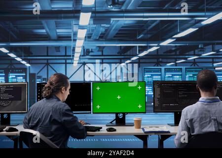 Colleagues working on green screen desktop PC in data center housing storage infrastructure. Coworkers using mockup computer display in server hub workspace filled with networking tech Stock Photo