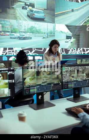 Team leader overseeing diverse operators in delivery call center office, employees using global positioning system to guide shipment trucks on city routes via satellite map. CCTV surveillance. Stock Photo