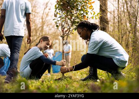 Two diverse girls working together to plant more seedlings and greenery, filling up ground holes and growing vegetation. Child and teenager collaborating on protecting the forest ecosystem. Stock Photo