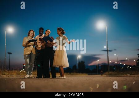 Group of diverse young adults collaborating outdoors under streetlights during dusk, showcasing teamwork and study in an urban environment with a Stock Photo