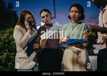 Team of professionals discussing documents outdoors during late evening meeting, with focused expressions, as they examine paperwork and engage in Stock Photo