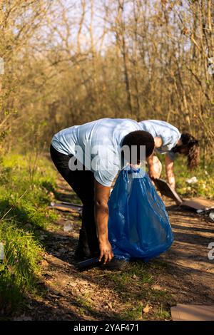 African american woman picking up rubbish and plastic waste from the forest, doing voluntary work to clear and preserve the nature. Volunteer using disposal bag to collect garbage junk. Stock Photo