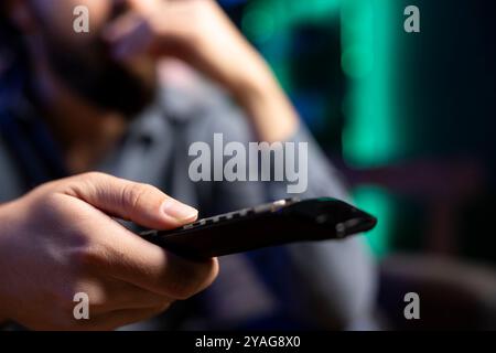 Focus on remote control held by man in blurry background enjoying leisure time at home watching TV. Close up shot of person chilling on couch in neon lit apartment, changing channels on television set Stock Photo
