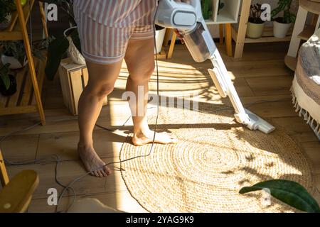 A woman vacuums a round carpet in a house among house plants with a hand vacuum cleaner. General cleaning of the house, cleaning service and housewife Stock Photo