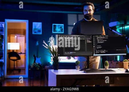 Cybersecurity expert using laptop to look for company security vulnerabilities, comparing with computer monitors. Programmer installing fortified code on laptop, crosschecking with PC screens Stock Photo
