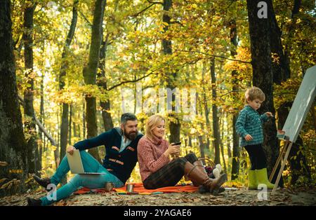 Unice son drawing his family giving joyful moments to mom and dad. Parents working using laptop and phone. Family picnic. Mother, father, son drawing Stock Photo