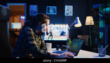 Smiling man satisfied about statistical data financial graphs on laptop screen while researching data. Remote worker happy about business graphic economic numbers, feeling pleased, camera B Stock Photo