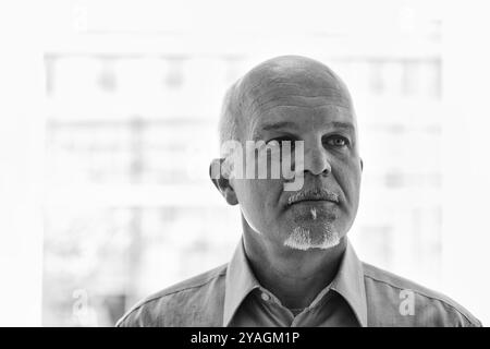 Mature male it specialist with a goatee is gazing thoughtfully through a window in an office building. The black and white image conveys a sense of ex Stock Photo