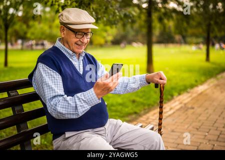Portrait of happy senior man in park. He is sitting on bench and using smartphone. Stock Photo