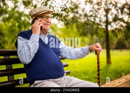 Portrait of happy senior man in park. He is sitting on bench and talking on phone. Stock Photo