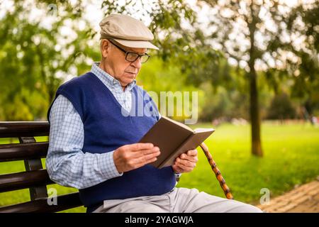 Portrait of senior man in park. He is sitting on bench and reading a book. Stock Photo